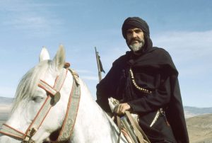 Sean Connery en chef berbère tout de noir vêtu pose fièrement sur un cheval blanc au coeur du désert dans le film Le lion et le vent.