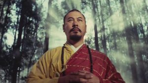 Un moine combattant les yeux dans l'horizon, concentré ; derrière lui une lueur blanche de soleil intense passe entre les arbres de la forêt ; scène du film A touch of zen.