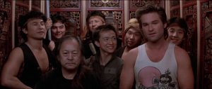 Kurt Russell entouré d'un groupe d'asiatiques dans une salle décorée à l'orientale, scène du film Les aventures de Jack Burton dans les griffes du mandarin.