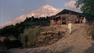Une silhouette d'homme vêtue de blanc monte la petite allée vers une maison de bois isolée au pied d'une montagne, scène du film Au-dessus du volcan.