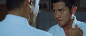 Deux judokas se font face, l'un est de dos, tenant celui d'en face par le col du kimono ; le judoka vu de face regarde son adversaire avec détermination et colère, scène du film Judo.
