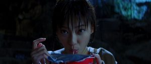Mona aspire une nouille d'un bol, debout, en regardant droit à la caméra dans le film Judo.