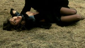 Une femme est étranglée, allongée sur l'herbe, par un homme tout vêtu de noir, scène du film Le couteau sous la gorge.