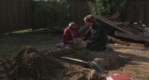 Les deux amis Glen et Terry dans le jardin, au soleil ; assis près du trou qu'ils ont creusé, ils inspectent la boule-météorite qu'ils prévoient d'enterrer, scène du film The gate.