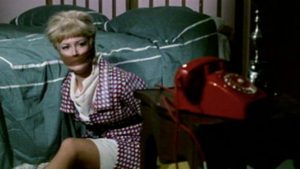 Au pied d'un lit, une femme attachée et bâillonnée regarde avec espoir un téléphone rouge, à quelque mètres d'elle ; scène du film Des fleurs pour un espion.