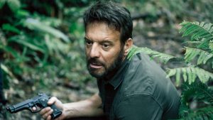 Dans la jungle, accroupi, Samuel Le Bihan est sur ses gardes, un revolver dans la main droite ; scène de la mini-série Ils étaient dix.