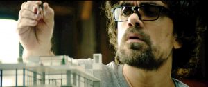 Peter Dinklage, concentré avec des lunettes sur le nez, met la dernière pièce à une maquette de bâtiment toute blanche dans le film Rememory.