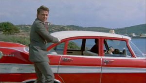 Alors qu'il s'apprête à monter à l'arrière de sa superbe Plymouth rouge et blanche, un homme se retourne brusquement, comme si on venait de lui faire peur ; scène du film Des fleurs pour un espion.