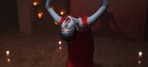 Une femme avec la peau bleue et une robe rouge hurle debout au milieu de quatre bougies ; elle est toute courbée comme si elle était possédée par un démon ; scène du film The Cursed Lesson.