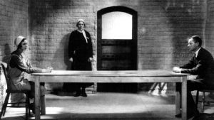 Plan d'ensemble sur une vaste cellule de prison, en pierre ; un homme et une femme se font face d'un bout à l'autre d'une table exagérment longue ; scène du film Meurtre de 1930.