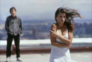Penelope Cruz sur un toit en ville, les cheveux au vents, se couvre avec ses bras laissant penser qu'elle a froid ; derrière elle la silhouette d'un homme dans le second plan flou ; plan du film Ouvre les yeux.