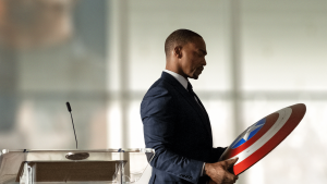 Anthony Mackie, en costume-cravate près d'un pupitre, tient le bouclier de Captain America dans ses mains et le regarde pensif ; scène de la série Falcon et le soldat de l'hiver.