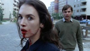 En pleine rue, au premier plan le visage d'Isabelle Adjani possédée, du sang lui coule de la bouche ; derrière elle Sam Neill s'approche d'elle, le regard presque lubrique ; scène du film Possession.