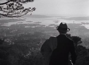 Raimu vu de dos en costume de ville et chapeau, une veste posée sur l'épaule, est debout sur une colline qui lui donne une vue sur Toulon et la mer dans le film L'étrange Monsieur Victor.
