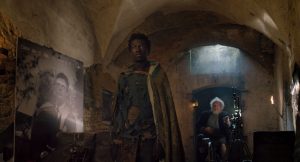 L'acteur Kalipha Touray est dans une crypte, vêtu comme un homme du Moyen-Âge, à l'arrière-plan un vieil homme assis sur un trône ; scène du film Last Words.