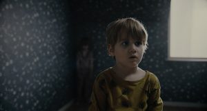 Dans sa chambre, un petit garçon avec un t-shirt jaune ne voit pas le fantôme d'enfant derrière lui, caché dans un angle de la pièce ; scène du film The Other Side