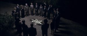 Un cercle d'hommes et de femmes en costume de ville noir est autour d'un pentagramme formé par des bougies sur le sol dans le film Starry Eyes.