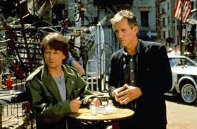 Mihael J. Fox et James Woods sont debout à la table haute d'un café en extérieur, ils regardent à l'horizon avec l'air interrogateur dans le film La manière forte.