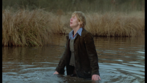Mimsy Farmer les yeux clos, en grande douleur, plongée toute habillée dans un cours d'eau qui lui monte jusqu'aux hanches ; plan iconique du film La Traque.