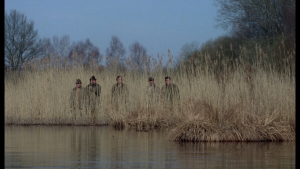 La silhouette de quatre chasseurs se dessine dans les hautes herbes, au bord d'un cours d'eau ; plan du film La Traque.