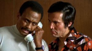 Woody Strode est au téléphone, Henry Silva est à ses côtés, tend l'eoreille pour entendre lui aussi ; scène du film Passeports pour deux tueurs de la Trilogie du Milieu.