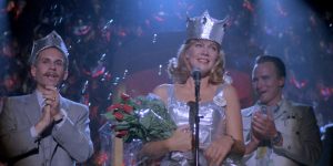 Une femme blonde en robe de cérémonie parle à un micro, tenant un bouquet et portant une couronne sur la tête ; à côté d'elle deux hommes l'applaudissent ; scène du film Peggy Sue s'est mariée de Francis Ford Coppola.