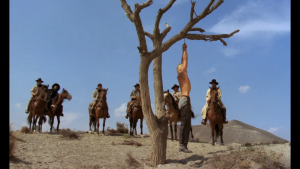 Un homme est pendu à un arbre par les mains, au beau milieu d'une plaine désertique, autour de lui six voyous à cheval le regardent ; scène du film Bianco Apache.