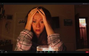 Capture d'écran d'ordinateur issue du film Host : une jeune femme assise à son bureau dans sa chambre sombre, les rideaux tirés, tient son visage entre ses mains, angoissée.