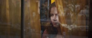 Amy Adams renfrognée derrière une fenêtre, entre deux rideaux couleur moutarde dans le film La Femme à la Fenêtre.