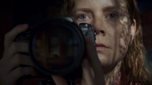 Gros plan sur le visage d'Amy Adams les cheveux mouillés, derrière un appareil photographique ; son regard n'est pas dirigé vers l'écran de contrôle de l'appareil mais vers l'horizon ; plan issu du film La Femme à la Fenêtre.