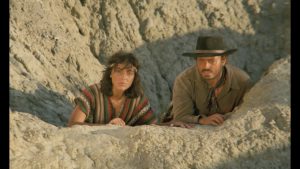 L'actrice Mapi Galan, en Amerindienne, et l'acteur Vassili Karis en cow-boy se cachent derrière une roche montagneuse, sous un soleil lourd dans le film Scalps. 