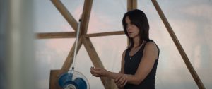 En plein air, debout près d'une structure en bois et d'un ventilateur, une jeune femme se tient le poignet, comme après une piqûre, et regarde devant elle l'air interrogateur ; scène du film La Nuée de Just Philippot.