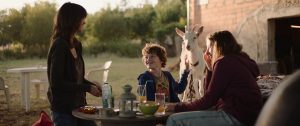 Scène de repas sur une terrasse en fin de journée, deux femmes face à face et un petit garçon entre elles, tout sourire, tenant une chèvre par le collier ; scène du film La Nuée de Just Philippot.