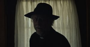 Devant une fenêtre aux rideaux blancs, un prêtre portant un chapeau nous regarde, avec un sourire inquiétant dans le film The Dark and the Wicked.