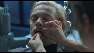 Plan du film Canine Panique de Baptiste Drapeau ou un homme est assis sur un siège de dentiste, les yeux clos, pendant qu'un autre homme s'occupe de ses dents, le tout baigné dans une lumière de nuit.