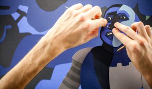 Deux mains modèlent le visage d'un bonhomme en papier, bleu et gris : ce sont les mains de Baptiste Drapeau.