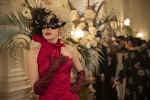 Cruella en belle robe de soirée rouge, portant un masque vénitien, porte la main à sa poitrine, surprise par quelque chose.