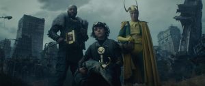 Trois hommes en costume de super-héros dont Lokin en armure divine, posent devant une ville grise et dont les bâtiments détruits.