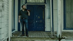 Jeremy Gardner se tient sur le palier d'une maison délabrée, un fusil à pompe posé sur l'épaule ; scène du film After Midnight.