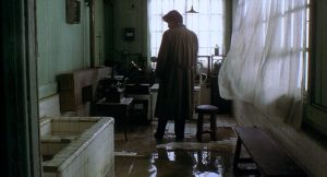 Une silhouette d'homme avec un long manteau est vue de dos, dans une salle de bains sinistre et sombre, avec de l'eau stagnante sur le carrelage ; plan issu du film Cure.