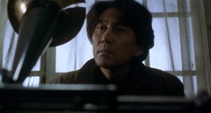 Le regard perdu de l'inspecteur Takabe se pose sur un vieux gramophone dans le film Cure.