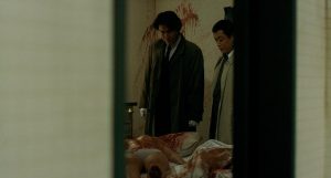 Vus dans l'embrasure d'une porte, deux inspecteurs en civil observent des corps ensanglantés à leurs pieds, cachés sous des draps blancs ; scène du film Cure.
