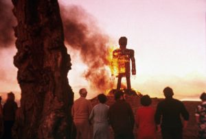 Un groupe d'hommes et de femmes vus de dos contemple une haute statue de bois prendre feu sous un ciel de crépuscule, scène du film The Wicker Man.