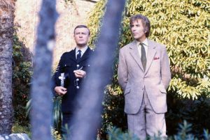 Dans le film The Wicker Man, un policer et un homme en costume patientent en plein air, derrière un arbre fin.