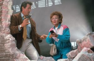Stallone, un revolver à la main est à côté d'une vieille dame en jogging bleu et rose ; tous deux sont à côté d'un mur en briques brisé dans le film Arrête ou ma mère va tirer !