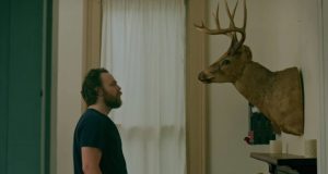 Un homme observe un buste de cerf empaillé, les bras le long du corps, dans un salon du film After Midnight.