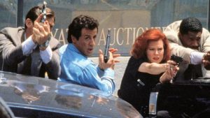 Quatre policiers dont Sylvester Stallone se protègent derrière le capot de voitures, armes à la main ; scène de fusillade dans le film Arrête où ma mère va tirer !