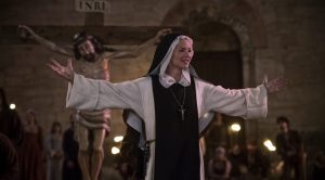 Benedetta (Virginie Effira) les bras en croix devant une statue de Jésus s'adresse à une petite foule dans la rue.