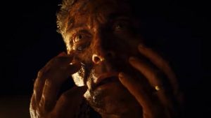 Dans un clair-obscur très marqué, un homme dont le visage semble en partie brûlé porte ses doigts à ses joues dans le film Old.