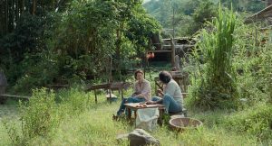 Elkin Diaz et Tilda Swintont discutent, assis près d'une table de pique-nique, dans un paysage vert de montagnes ; plan issu du film Memoria.